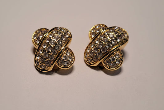 A pair of gold-toned statement earrings covered in clear pavé stones, displayed on a light gray surface.
