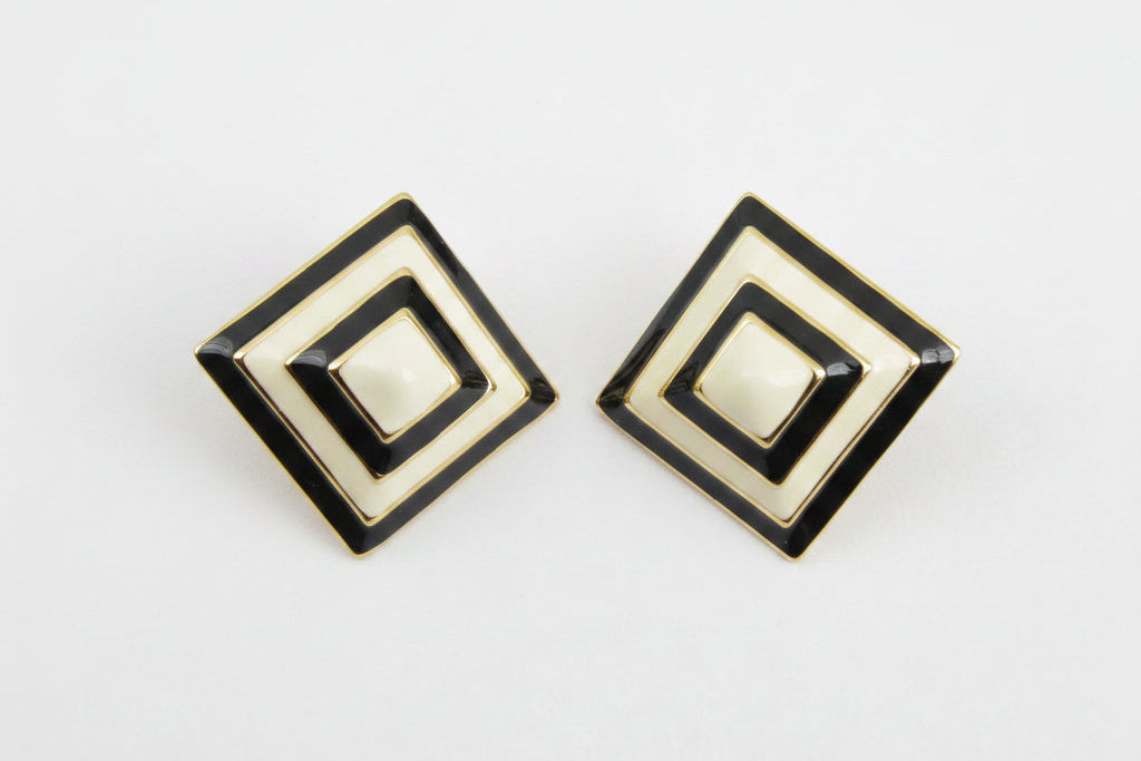 A pair of square geometric stud earrings with concentric diamond designs in cream, black, and gold, displayed on a neutral light gray background.
