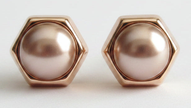 A pair of hexagonal stud earrings featuring lustrous rose gold-toned metal frames and smooth, pearl-like oval centers in a soft champagne pink hue, set against a clean white background.