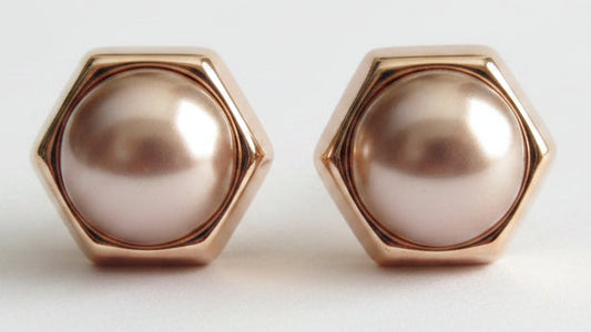 A pair of hexagonal stud earrings featuring lustrous rose gold-toned metal frames and smooth, pearl-like oval centers in a soft champagne pink hue, set against a clean white background.